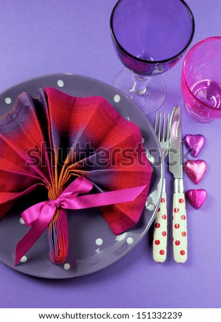 Pink and purple table setting with fan shape napkin and polka dot plate and cutlery with heart shape chocolates and pink & purple glasses for fun birthday or special occasion dining table. Vertical.