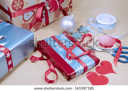 Christmas gift wrapping with red, white and aqua blue wrapping, hearts, tags, scissors and gift boxes with festive Christmastime ornament decorations on a pale wood table desk.  Selective focus.