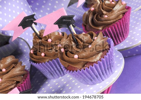 Graduation day pink and purple party chocolate cupcakes with small graduation cap toppers on polka dot stand - close up.