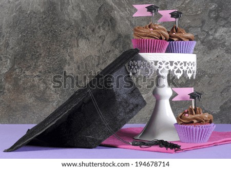 Graduation day pink and purple party chocolate cupcakes on white vintage white stand with graduation cap against stylish modern black slate background.