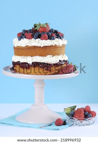 Fresh whipped cream and berries layer sponge cake on pink cake stand against pale blue and white background.