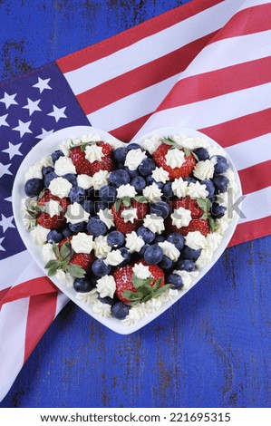 Patriotic red, white and blue berries with fresh cream stars in white heart shape flag on blue wood background, with American USA stars and stripes flag, for Independence Day or Columbus Day party .
