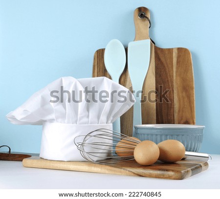 Modern kitchen cooking kitchenware and chef\'s hat with mixing bowl, whisk, chopping boards and eggs on a pale aqua blue and white background.