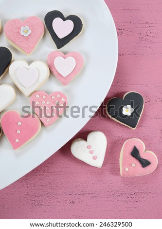 Romantic heart shape pink, white and black cookies on white heart shape plate on vintage shabby chic pink wood table for Valentine or wedding day, vertical overhead.