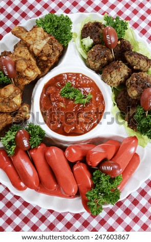 Super Bowl Sunday football party celebration food platter with chicken buffalo wings, meat balls, hot dogs and salsa dip on red check table cloth, vertical overhead.
