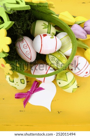 Happy Easter green and yellow felt basket of of pink, white and green easter eggs on rustic vintage yellow wood table, with heart shape greeting card, vertical.