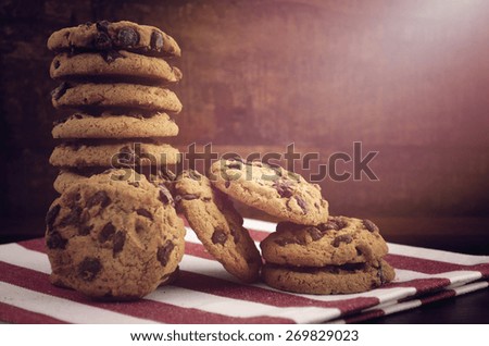 Stack of chocolate chip cookies on red and white stripe napkin against a dark wood background, with applied vintage style filters and added light stream.