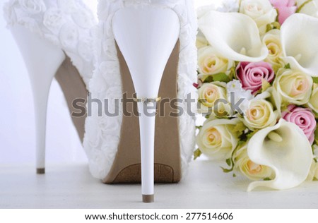 Wedding Day concept with rings on heel of beautiful white floral bridal stiletto shoes with bouquet on white shabby chic wood table.