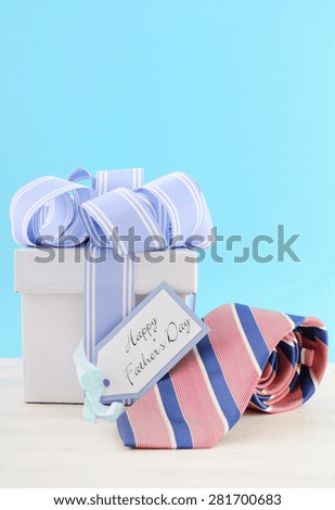 Happy Fathers Day Gift with Blue and White Ribbon with red and blue stripe neck tie and gift tag on white wood table and pale aqua blue background.