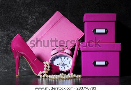 Ladies pretty pink female office desk with high heel shoe, clock, pen, stationery filing boses, pearls and diary organizer against a dramatic black background.