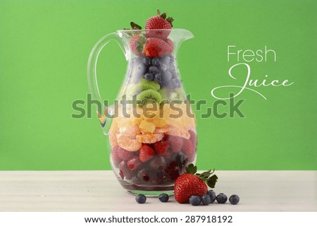 Fresh fruit juice concept with rainbow color fruit including cherries, strawberries, raspberries, mandarin segments, kiwi fruit, and blueberries in tall glass jug, against  modern green background.