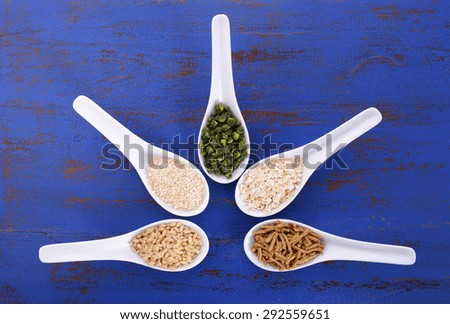 Healthy High Fiber Prebiotic Grains in serving spoons, including wheat bran cereal, oat flakes, dried legume peas, oat bran and pearl barley, on rustic dark blue wood table.