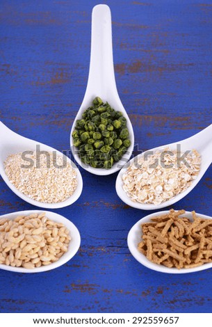 Healthy High Fiber Prebiotic Grains in serving spoons, including wheat bran cereal, oat flakes, dried legume peas, oat bran and pearl barley, on rustic dark blue wood table.