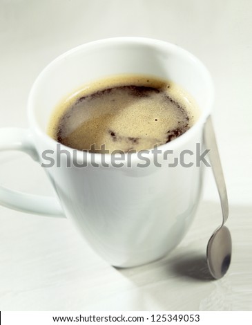 High angle view of a mug of rich strong American coffee topped with froth in a plain white ceramic mug with a silver serving spoon