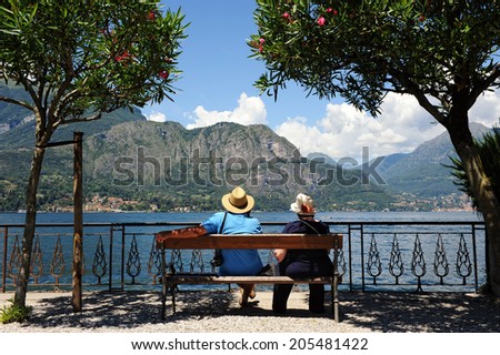 Senior tourist couple sitting on a bench in front of an ornate wrought iron railing admiring a beautiful scenic mountain lake, view from behind