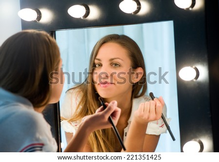 Attractive young woman applying her makeup reflected in a mirror with a light bulb surround looking at the camera with a friendly smile