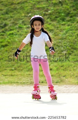 Little girl roller skating on roller blades in her safety helmet and gloves approaching the camera on a rural road with a happy smile