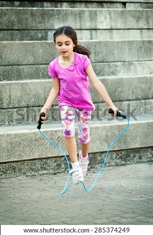 Happy attractive little girl in a trendy pink outfit skipping outdoors using a skipping rope at the bottom of a flight of steps
