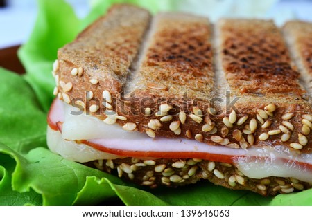 Integral toast with sesame and other whole seeds, ham and cheese sandwich served on a brown porcelain plate over fresh lettuce leaf, close up with shallow depth of field