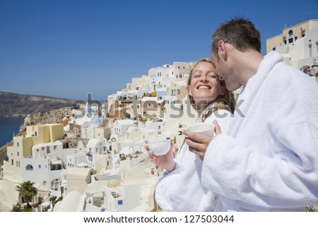 Couple in bathrobes with cups smiling with scenic mediterranean village in the background
