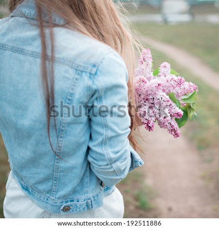 Picture from behind girl in a denim jacket and lilac flowers in their hands