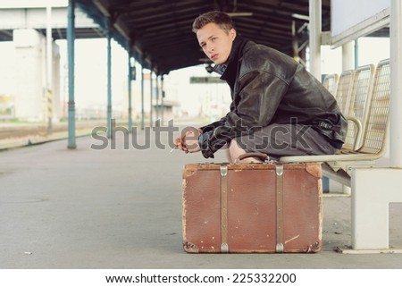 Stylish guy sitting at the station with a cigarette and an old suitcase