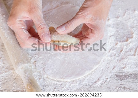 Woman hands making dumplings, closeup, from above view. Handmade dough and dough roller are on wooden kitchen counter with flour