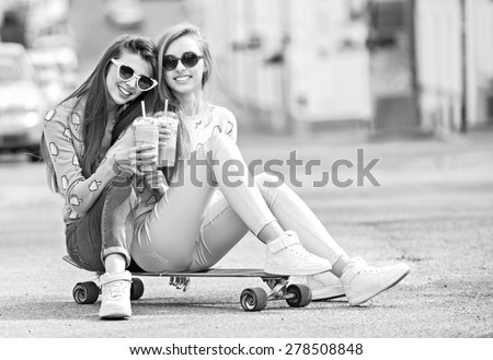 Beautiful young girls hipster girlfriends posing with a skateboard seat on skate, street fashion lifestyle in sunglasses. Keep cocktail and smiling