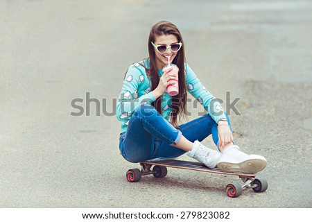 Beautiful young woman posing with a skateboard seat on skate, street fashion lifestyle. Keep cocktail and make selfie photo
