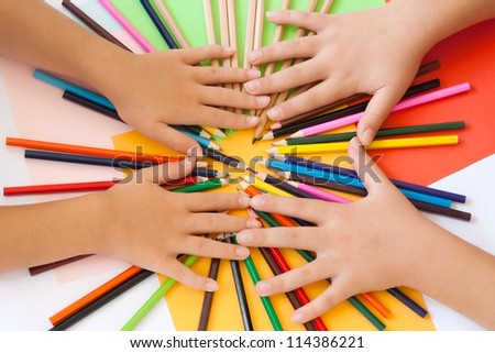 Wooden pencils and two pairs of children\'s hands from above. Paper in more colors in the background. Top view.