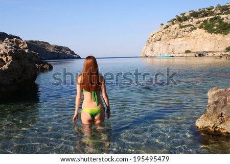 Young red haired woman in swimsuit standing back go to swim in beautiful bay clear water and looking to a sea . summer