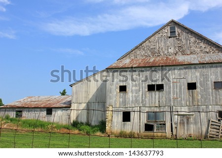 Old weathered gray barn with open windows and doors, fenced in with chicken wire out in rural countryside.