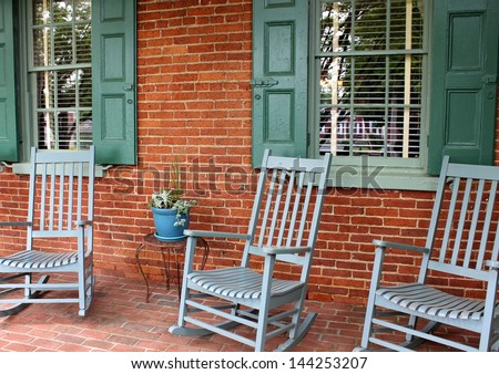 Inviting front porch with brick wall and floor, warm green shutters and trio of rocking chairs for company to sit and relax on ,