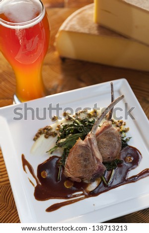 Grilled lamb chops on a white plate with a glass of beer and a block of round cheese.