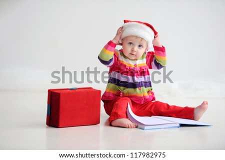 Baby sitting on the floor in a red santa hat. Kid holding his head. Christmas. On the floor is a book.