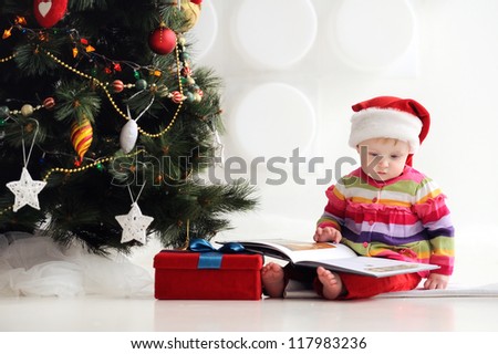 Baby sitting on the floor in a santa hat and reading a book. A child in a red cap. In the background, Christmas tree with ornaments. Christmas. The floor box with gifts.