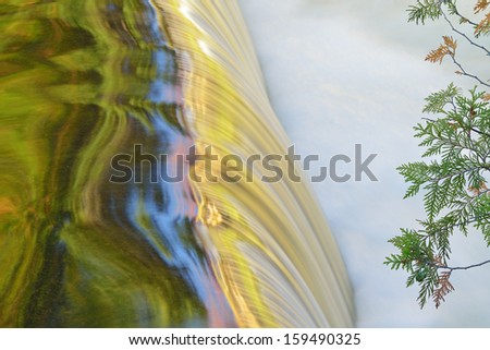 Bond Falls cascade framed by a cedar branch, captured with motion blur, and illuminated by reflected color from sunlit autumn trees and blue sky overhead, Michigan\'s Upper Peninsula, USA
