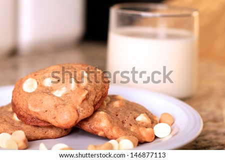 A plate of home made white chocolate and macadamia nut cookies are garnished with additional nuts and white chocolate chips and served with almond milk.