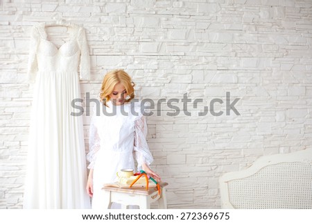 Beautiful blond bride near white brick wall inside light interior. Long wedding white dress on the wall and small table with gourmet food