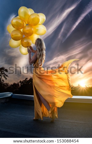 Very attractive young woman in beautiful yellow dress with yellow balloons standing on the roof.