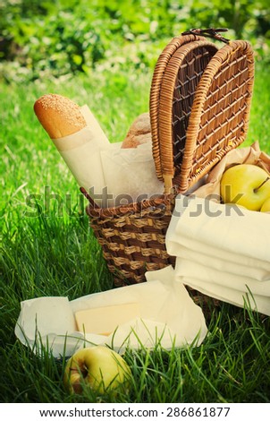 Picnic Wattled Basket on Green Grass with Fresh Bread, Apples, Cheese