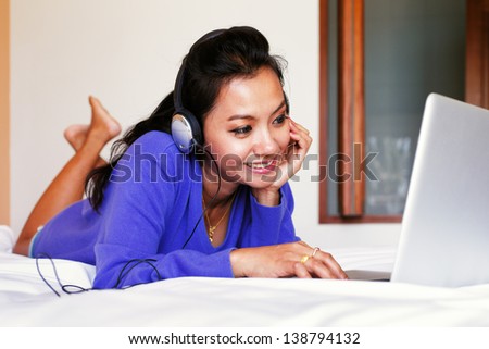 Young smiling woman in a blue top and headphones using a laptop in bed