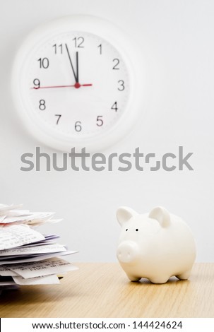 Tax time. A stack of bills next to a piggy bank with a clock behind