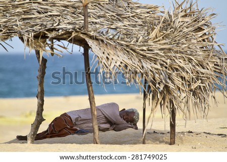 Batticaloa, Sri Lanka - June 24, 2014: Old tamil ethnicity man resting in shadow in Sri Lanka beach. Is making nap in palm leaves shadow.