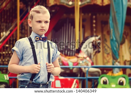 Sad boy near a game park attraction. A boy in a shirt with a stylish haircut and a butterfly. Boy waiting for a ticket to the roundabout. A boy wearing blue shirt and pants with suspenders.