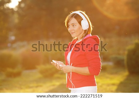 Woman lit by the rays of the evening sun at sunset. Good mood, favorite music, happy time. Woman standing listening to music with her mp3 player and headphones against nature. Woman urban clothing.