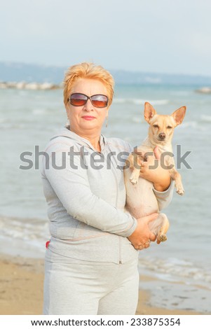 Adult woman is holding her dog. Retired woman standing on the shore of the sea or ocean. Old woman spends time with his dog on the background of the sea. Walking the dog on the beach. Tourism. Dog.