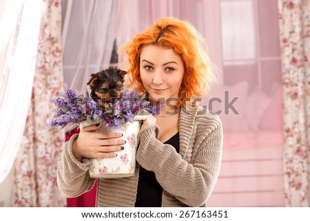 Red haired girl is holding a puppy dog and flowers. Young beautiful woman in home interior with a puppy breed York. Puppy sitting in a basket with flowers. Woman, comfort, home, happiness -  life joy.