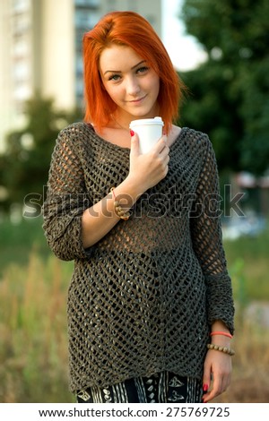 Red hair woman drinking tea or coffee from a white paper disposable cup. Concept - tea or coffee cup. Stretching forward with coffee cup. Young beautiful girl on the background of nature city park.