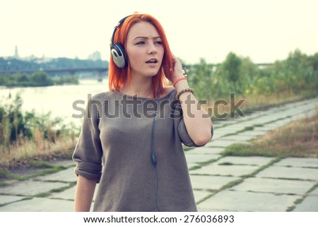 Young and beautiful woman wearing headphones listening to music and singing on the background of nature city park and river. Woman with headphones listening music. Music teenager girl dancing against.